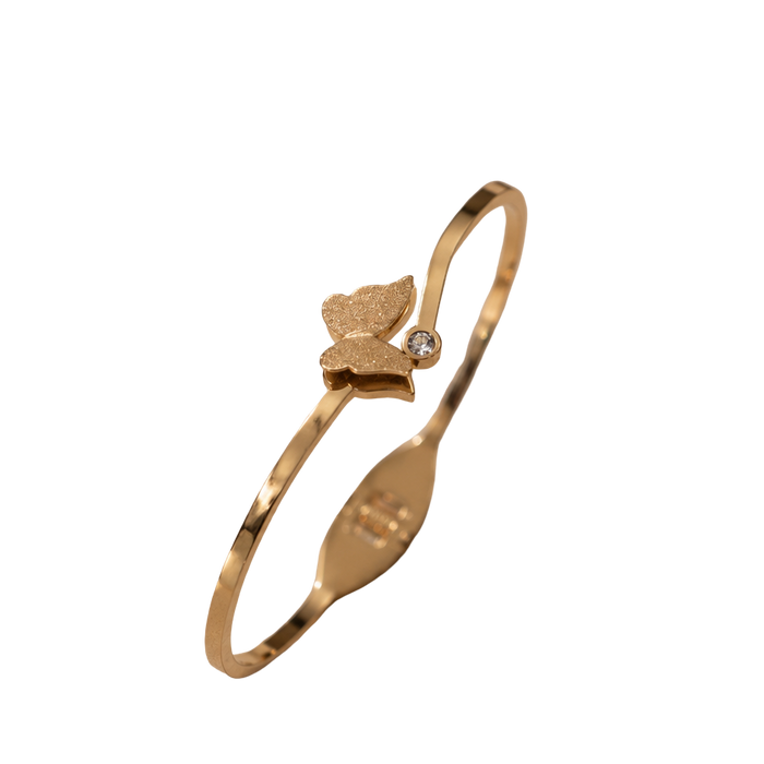 A gold-plated bangle bracelet with a small butterfly charm and a tiny clear stone, shown on a plain white background.