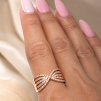 A close-up of a hand with pink manicured nails wearing a rose gold ring with multiple twisted bands set with small clear stones, against a soft beige background.