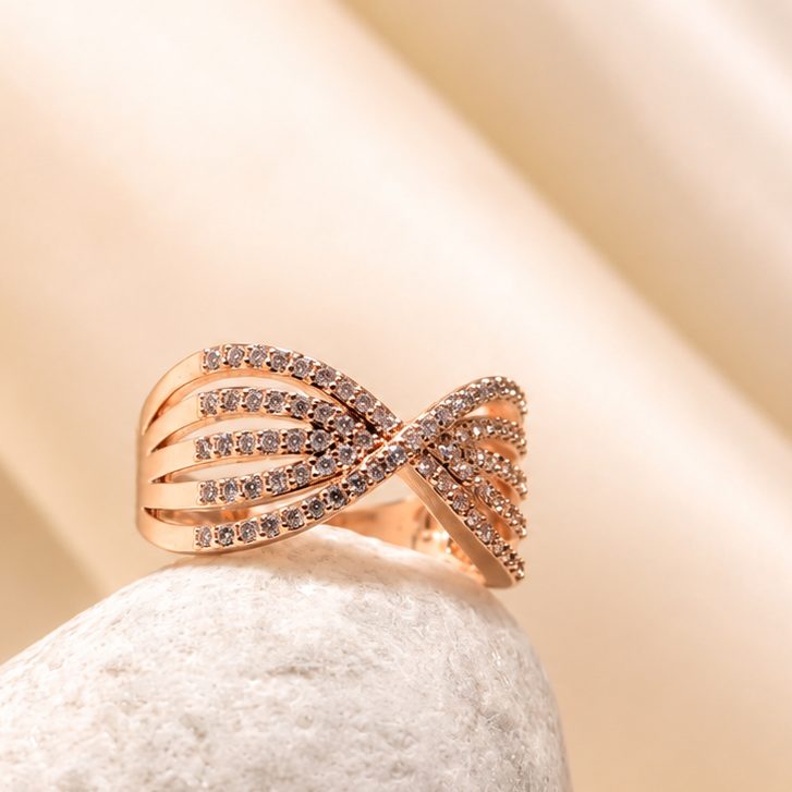 A rose gold ring with a twisted ribbon design set with small clear stones, resting on a textured white stone against a draped beige satin background.