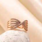 A rose gold ring with a twisted ribbon design set with small clear stones, resting on a textured white stone against a draped beige satin background.