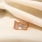 A rose gold ring with pavé-set clear stones in a twisted bow design, resting on draped cream-colored fabric.