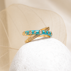 A gold ring with turquoise blue enamel and small clear stones, displayed on a white textured stone against a soft beige fabric background.