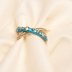 A close-up of a gold ring with turquoise blue inlay and small clear stones, displayed on draped cream-colored fabric.