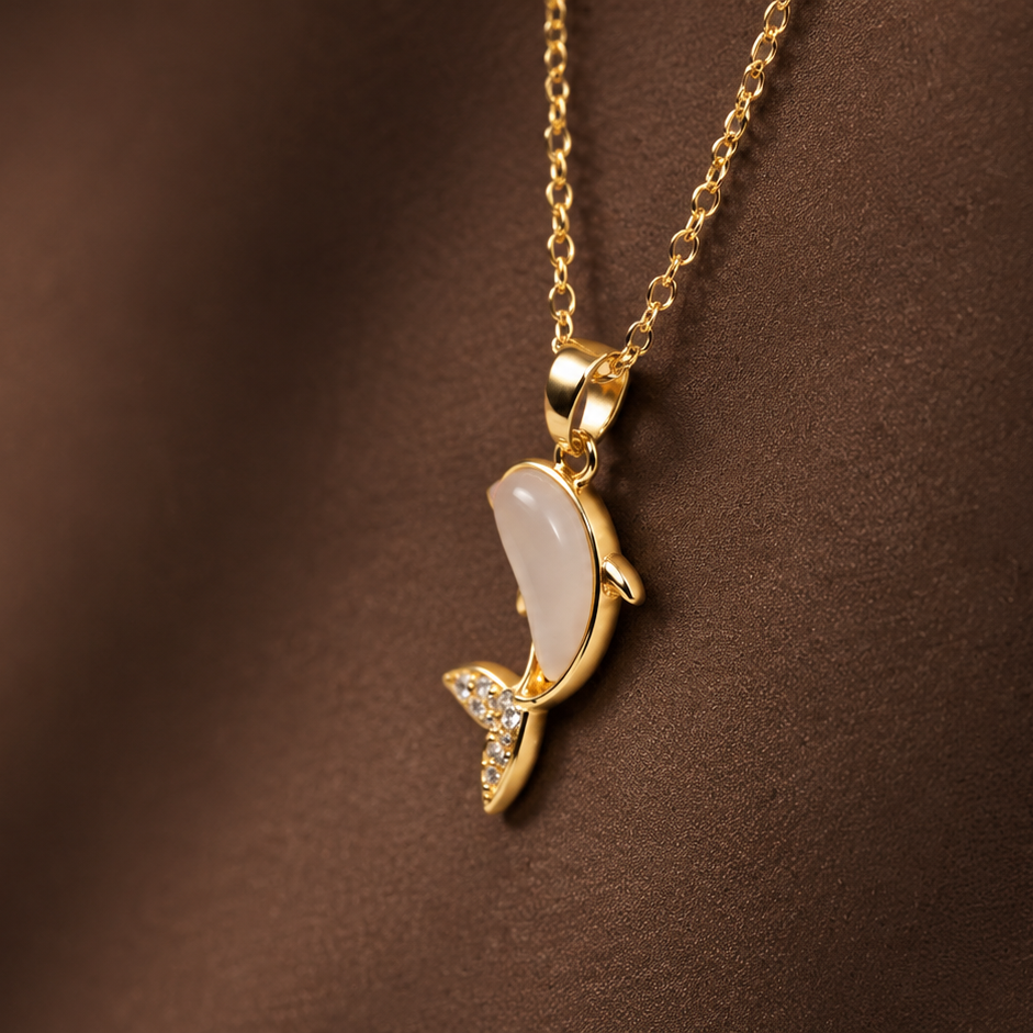 A gold pendant necklace featuring a stylized dolphin or fish-shaped charm with a pale iridescent center and small clear stones, shown against a dark brown textured background.