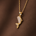 A gold pendant necklace featuring a stylized dolphin or fish-shaped charm with a pale iridescent center and small clear stones, shown against a dark brown textured background.