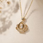 A gold crown pendant necklace with a clear teardrop-shaped center stone and small clear stones along the sides, displayed on a textured cream surface with soft shadows.