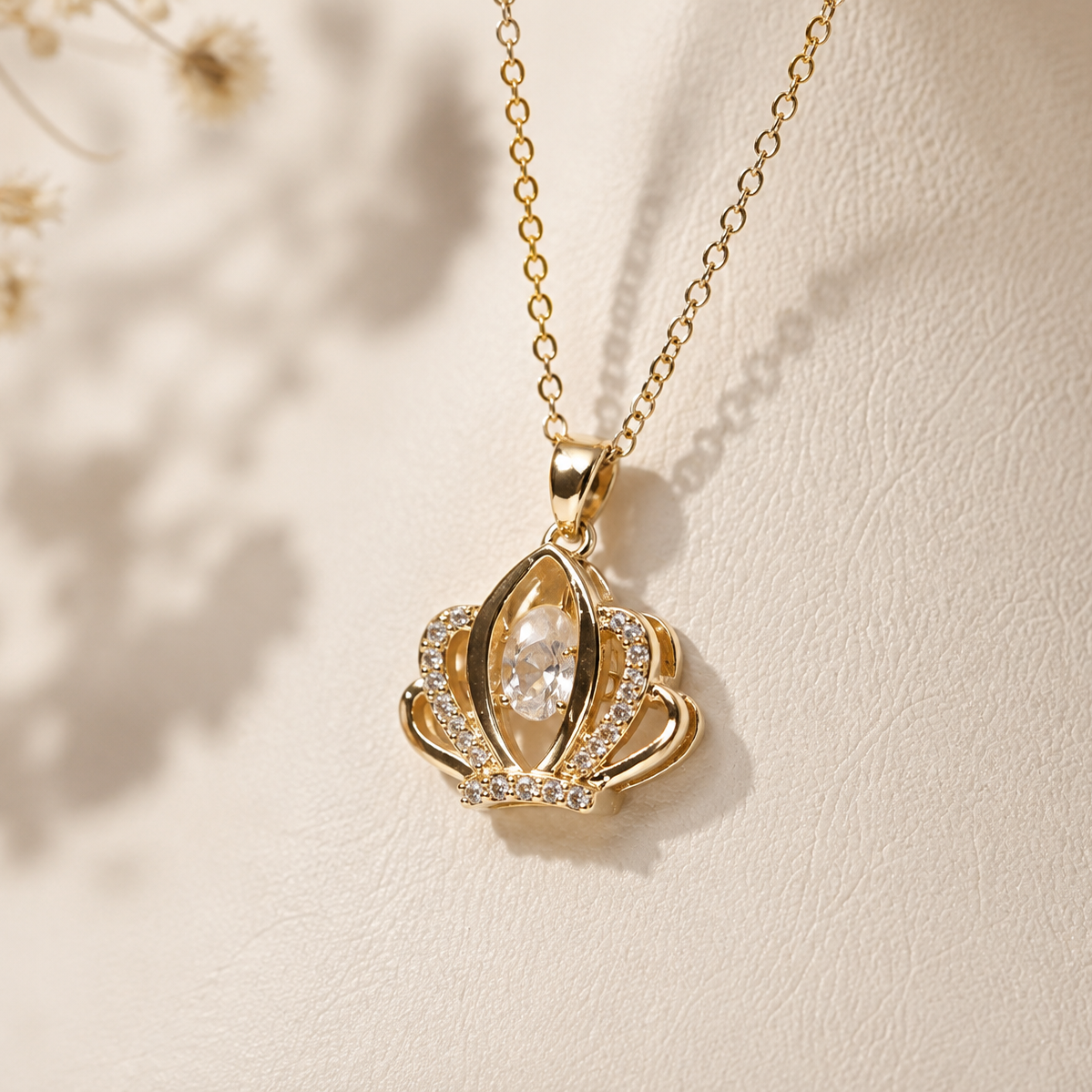 A gold crown pendant necklace with a clear teardrop-shaped center stone and small clear stones along the sides, displayed on a textured cream surface with soft shadows.