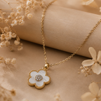 A gold-tone flower pendant necklace with a white inlay and a small clear stone center, displayed on a beige fabric surface with dried floral decorations.