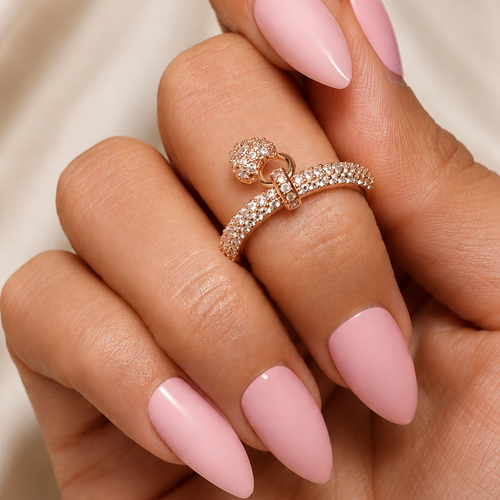 A close-up of a hand with long, glossy light pink manicured nails, wearing a delicate rose gold ring with pavé-set clear stones on the ring finger, set against a soft beige fabric background.