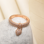 A rose gold ring with a pavé band and a small heart-shaped pendant, displayed on a textured gray stone against a soft beige background.