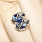 A close-up of a silver-toned ring featuring two heart-shaped deep blue gemstones accented with small clear crystals, resting on draped cream-colored fabric.