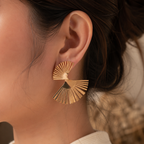 A close-up of a person's ear wearing a gold fan-shaped stud earring with radiating slits.