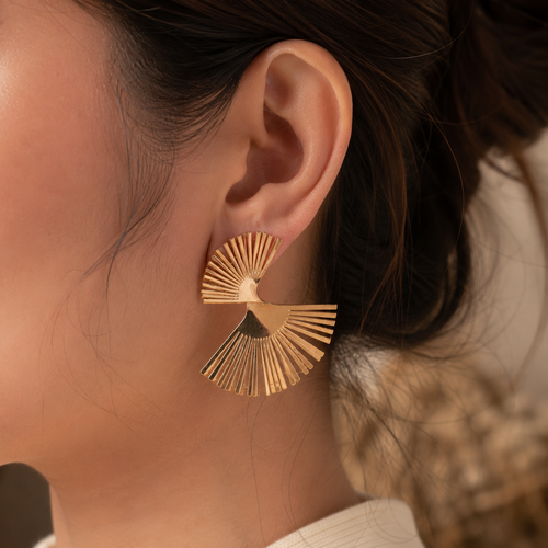 A close-up of a person's ear wearing a gold fan-shaped stud earring with radiating slits.