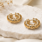 A pair of textured gold hoop earrings displayed on a white stone slab with soft beige fabric and dried flowers in the background.