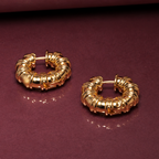 A pair of textured gold hoop earrings displayed on a dark burgundy surface.