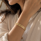 A close-up lifestyle shot of a woman wearing a slim gold bangle bracelet with engraved lettering, paired with a cream-colored blouse.