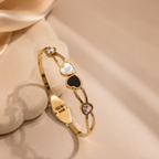 A gold-plated bangle bracelet with heart-shaped inlays (white, black, and pinkish-red) and small clear stones, displayed on a beige satin fabric background.