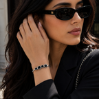 A woman wearing black cat-eye sunglasses and a black blazer, shown in a close-up outdoor shot. She is wearing a silver-toned chain bracelet with black rectangular links on her wrist.