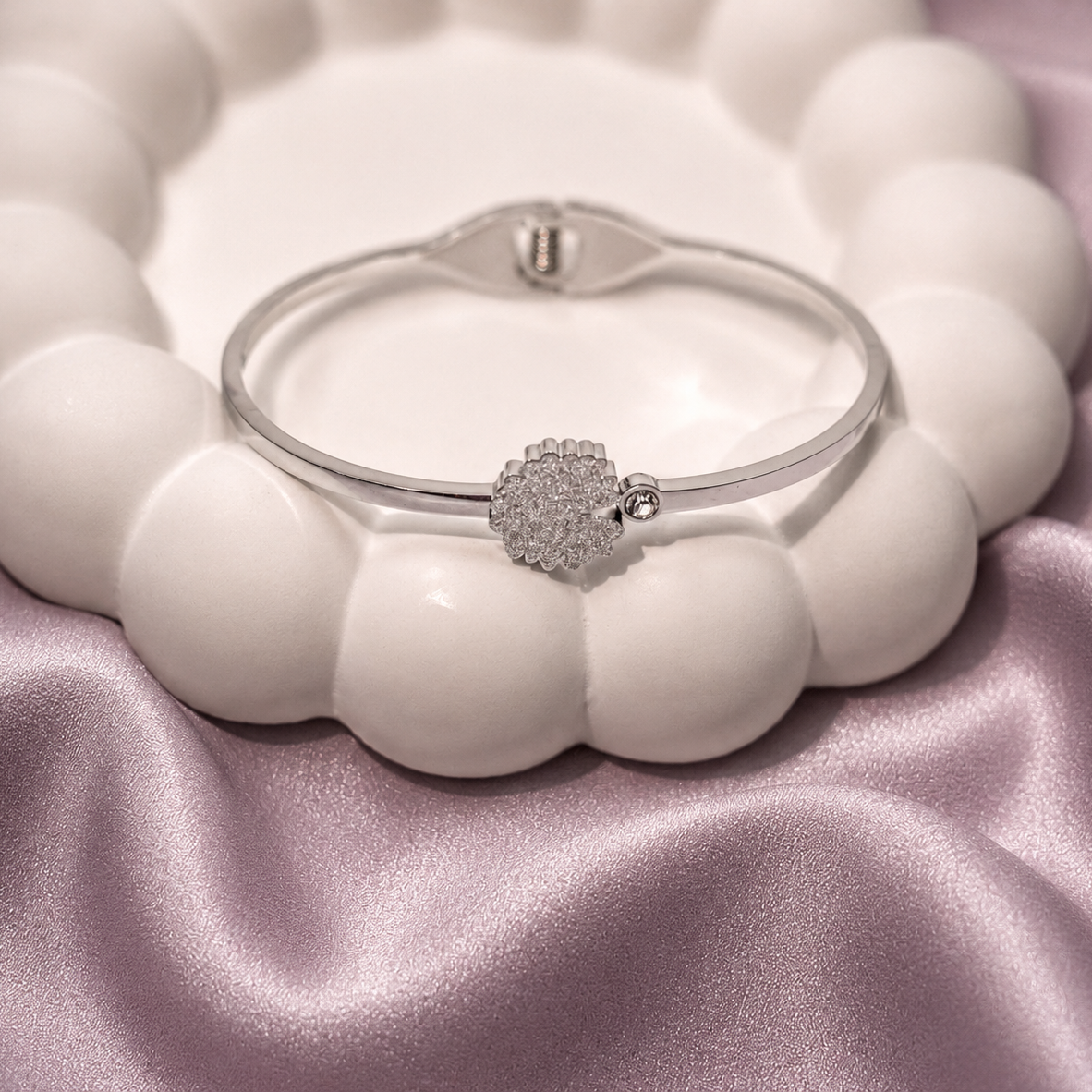 A silver-toned bangle bracelet with a pavé diamond heart charm, displayed on a white beaded stand over a pink satin fabric background.