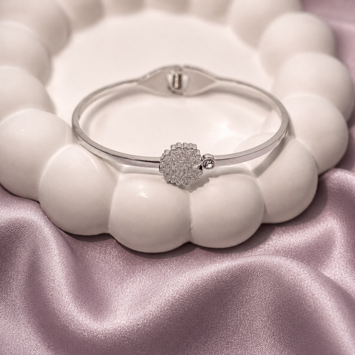 A silver-toned bangle bracelet with a pavé diamond heart charm, displayed on a white beaded stand over a pink satin fabric background.