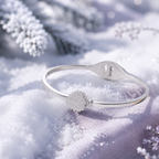 A silver-toned bangle bracelet with a textured snowflake charm, resting on a bed of white snow with blurred winter foliage in the background.