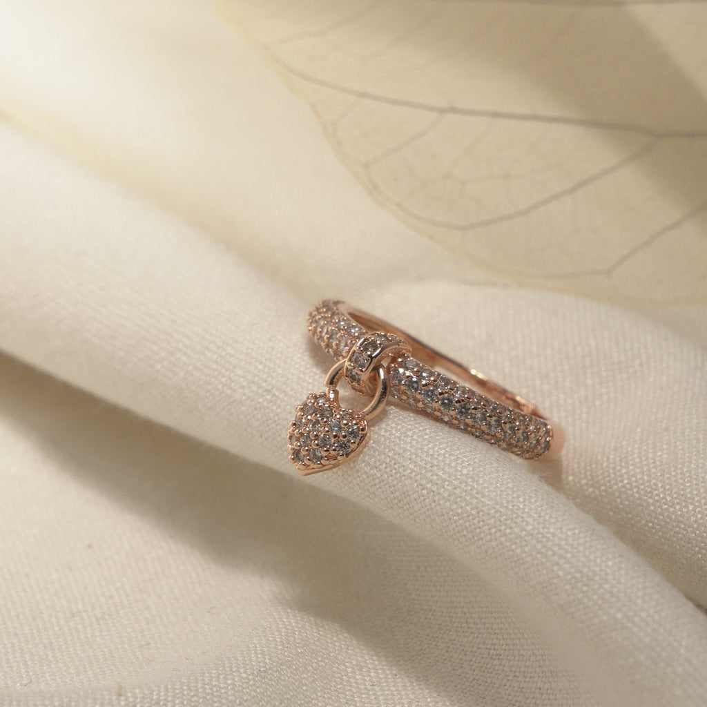 A delicate rose gold ring with a small heart-shaped pavé pendant, resting on soft cream-colored fabric with a translucent leaf overlay.