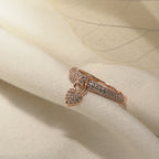 A delicate rose gold ring with a small heart-shaped pavé pendant, resting on soft cream-colored fabric with a translucent leaf overlay.
