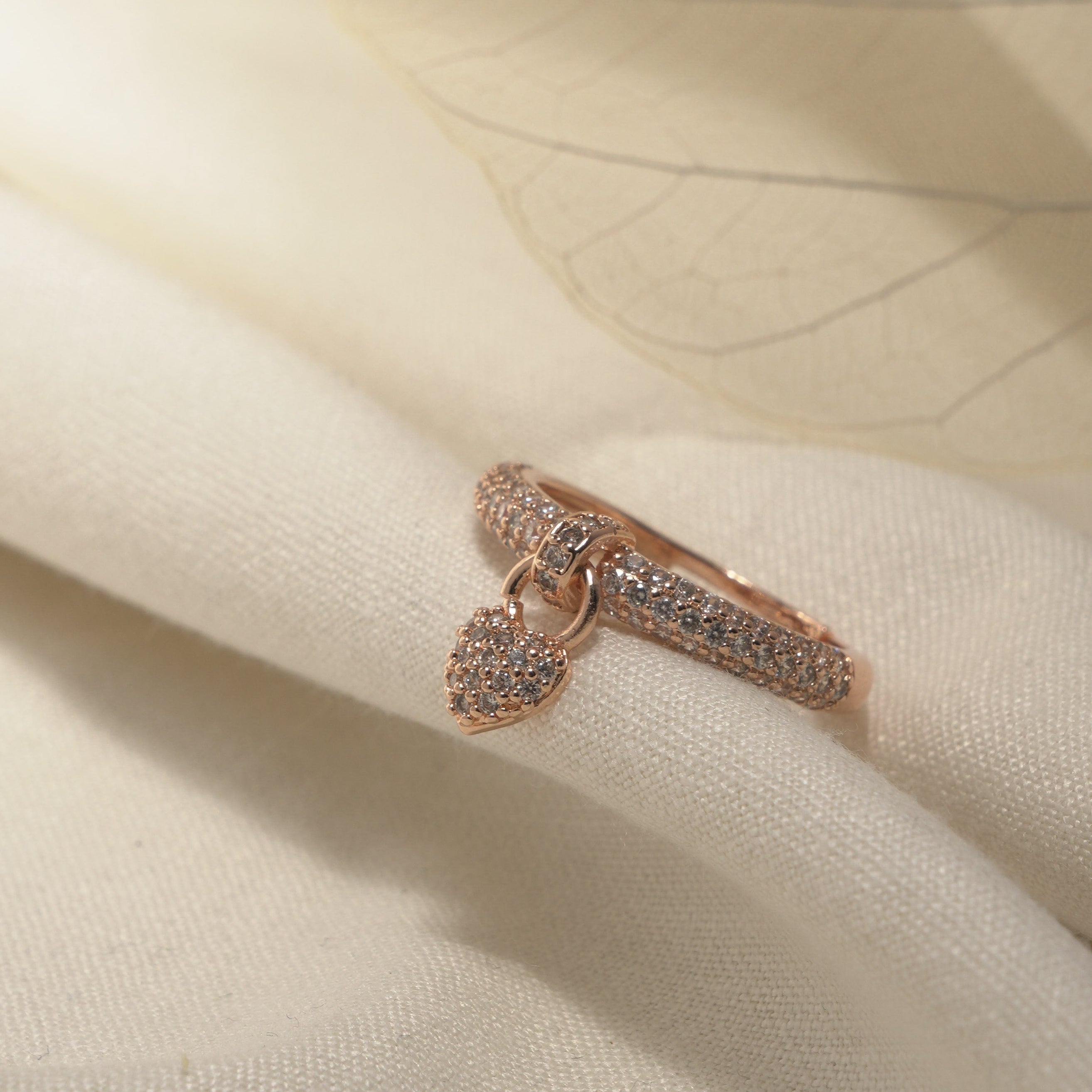 A delicate rose gold ring with a small heart-shaped pavé pendant, resting on soft cream-colored fabric with a translucent leaf overlay.