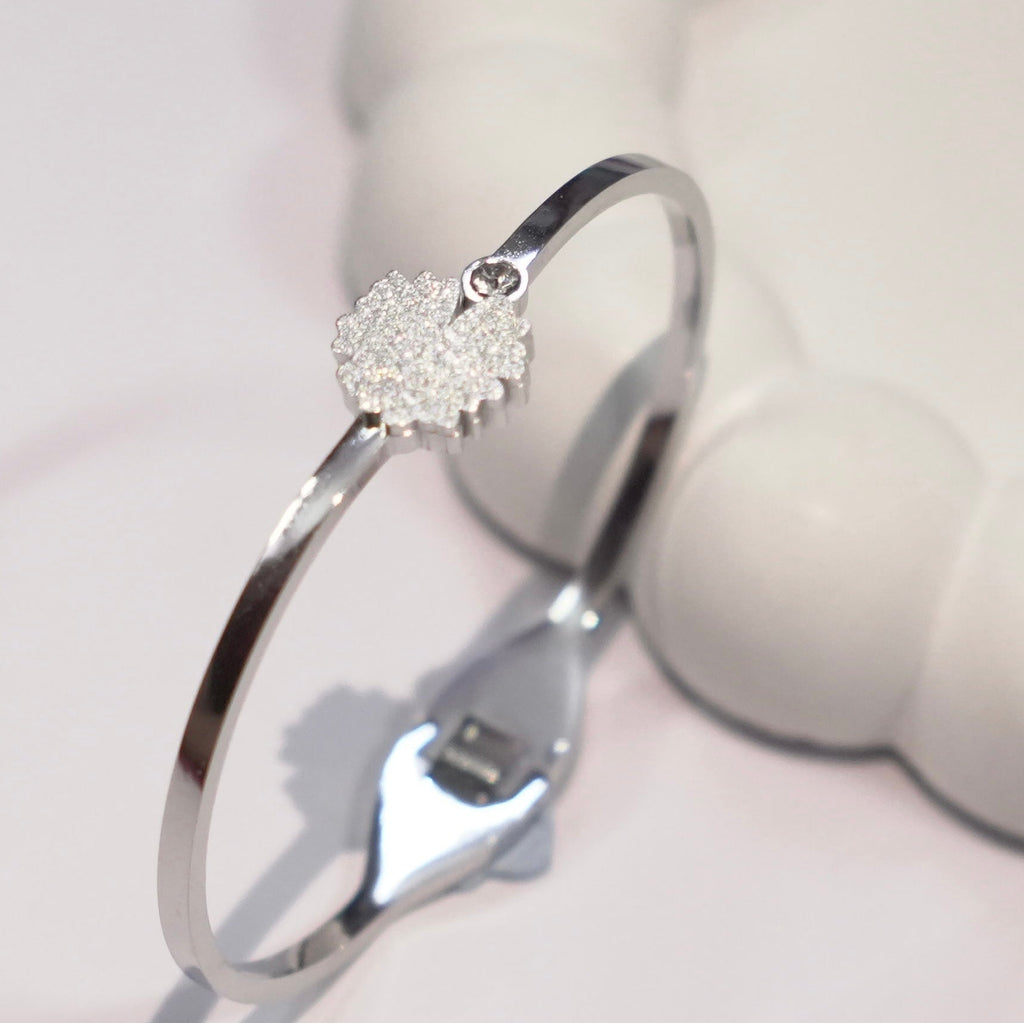 A polished silver-toned bangle bracelet with a small heart-shaped crystal cluster charm, displayed on a soft white surface.