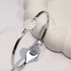 A polished silver-toned bangle bracelet with a small heart-shaped crystal cluster charm, displayed on a soft white surface.