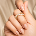 A close-up of a hand with manicured nails wearing two gold rings, one with small clear stones, against a soft beige fabric background.