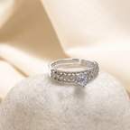 A silver-toned ring with a small clear center stone and pavé-set side stones, displayed on a textured stone pedestal against a draped cream satin background.
