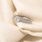 A silver-toned diamond ring with a central round diamond and a pavé band, resting on draped cream-colored satin fabric.