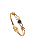 A gold-plated bangle bracelet featuring three heart-shaped inlays in white, black, and brown, with small clear stones at the ends, shown on a plain white background.