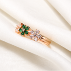 A rose gold ring with a floral design featuring green emerald-cut stones and clear round-cut stones, displayed on draped white satin fabric.