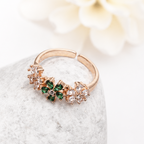 A rose gold ring with a floral design featuring green and clear crystals, resting on a textured gray stone with a soft white flower in the background.