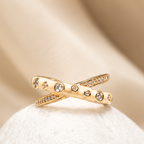A gold ring with a bow-like twisted design and small clear stones, displayed on a white stone against a soft beige fabric background.