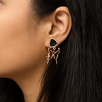 A close-up of a model's ear wearing a unique dangle earring. The earring features a triangular black stone top set in rose gold, with a dangling rose gold branch-like design.