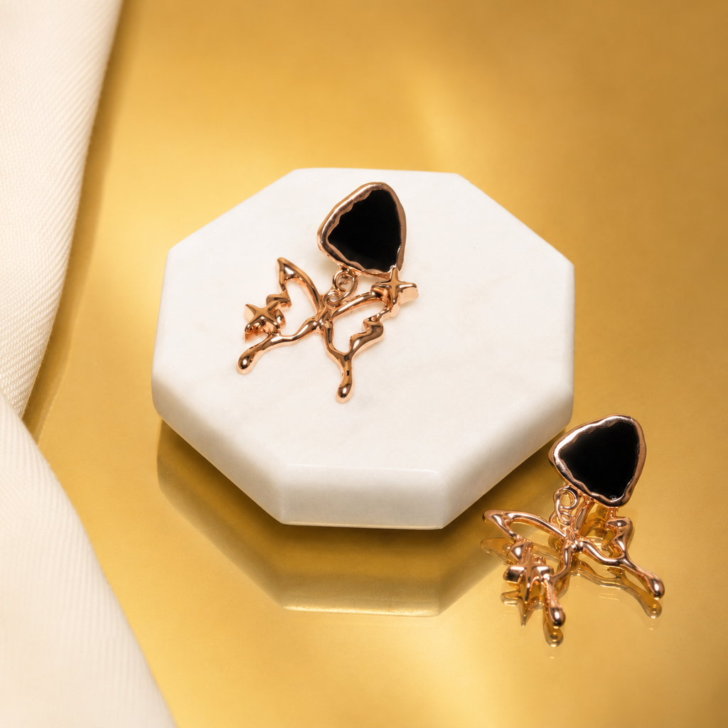 A pair of rose gold fairy earrings with black heart-shaped stones, displayed on a white octagonal stand against a metallic gold background.