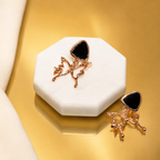 A pair of rose gold fairy earrings with black heart-shaped stones, displayed on a white octagonal stand against a metallic gold background.