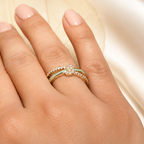 A close-up of a hand wearing two delicate gold rings, one with a small green stone and one with a floral cluster of clear stones, set against a soft cream satin background.