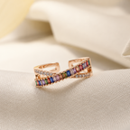 A rose gold open band ring featuring a central channel of colorful rectangular gemstones, resting on draped cream-colored fabric.