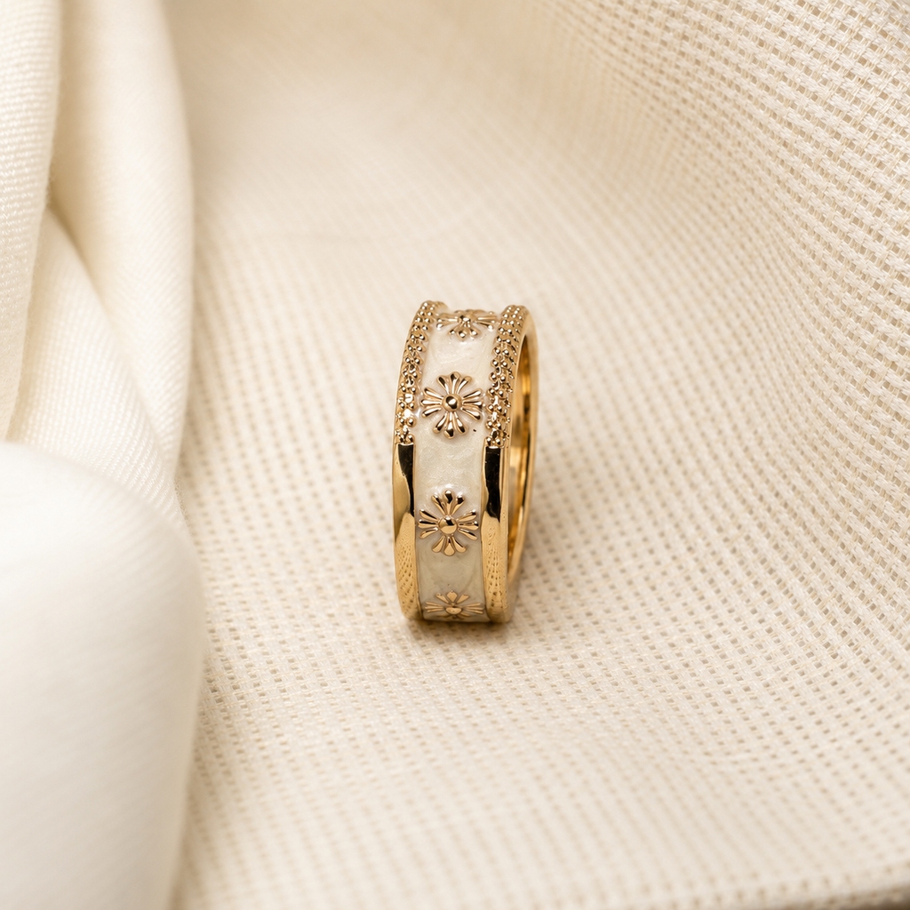 A gold ring with white enamel inlays and floral motifs, resting on a textured cream fabric background.