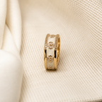 A gold ring with white enamel inlays and floral motifs, resting on a textured cream fabric background.