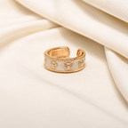 A gold-toned adjustable ring with a white enamel inlay and embossed floral star motifs, resting on draped cream-colored fabric.