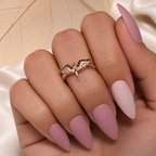 A close-up of a hand with long, almond-shaped matte pink nails, wearing a delicate rose gold ring with small black and clear stones on the ring finger, set against a soft beige and white background.
