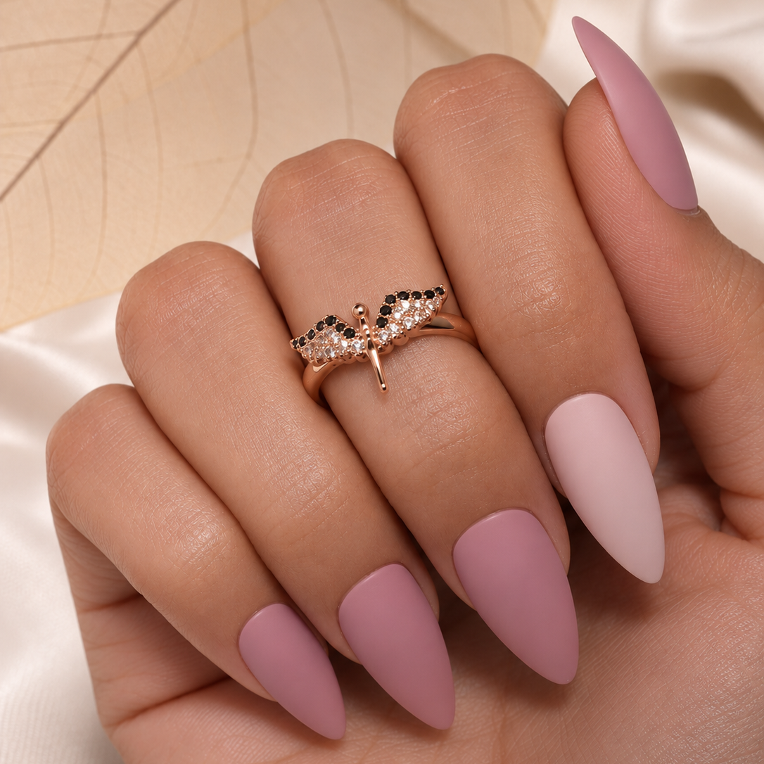 A close-up of a hand with long, almond-shaped matte pink nails, wearing a delicate rose gold ring with small black and clear stones on the ring finger, set against a soft beige and white background.